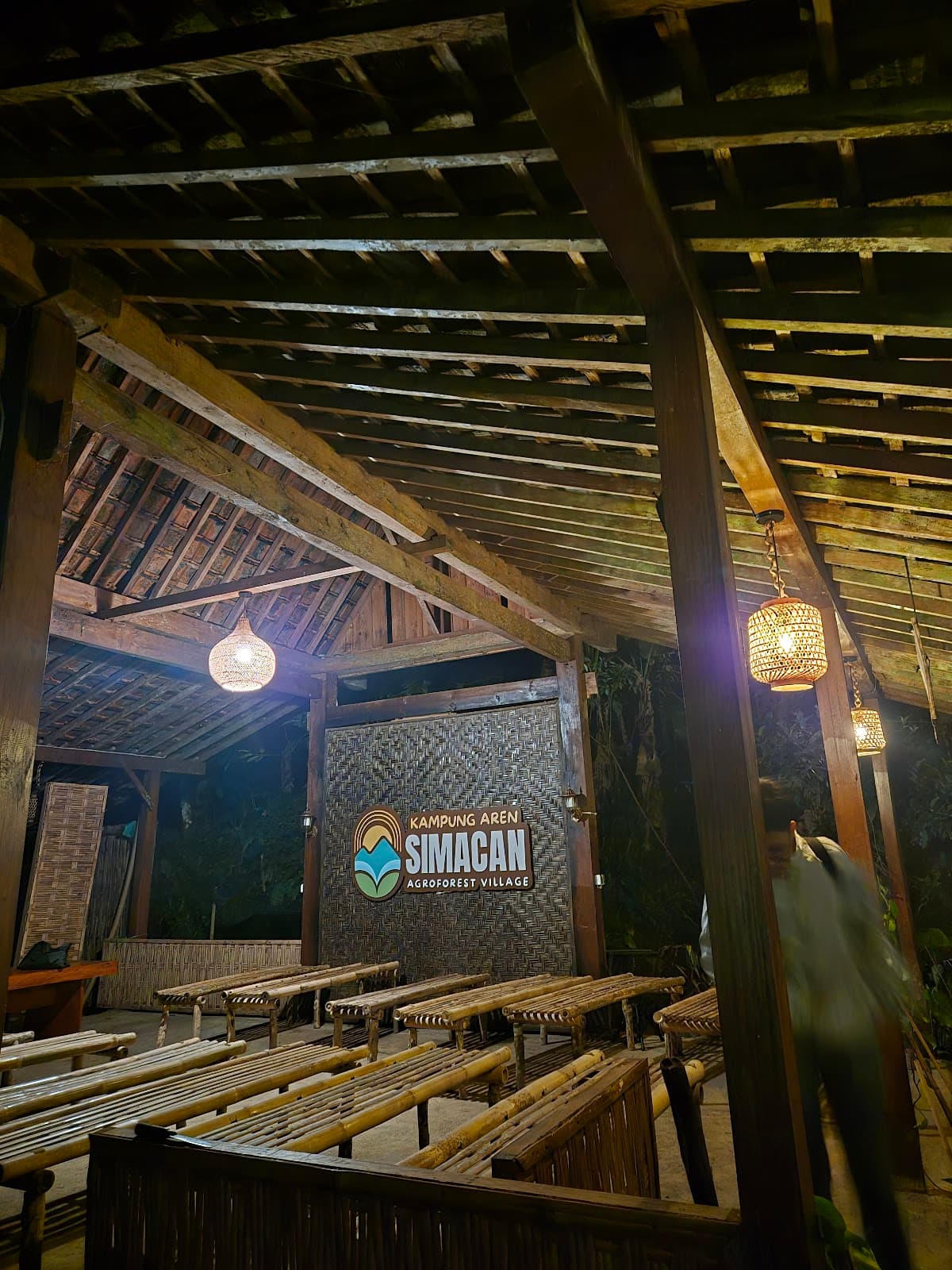 Interior balai bambu Kampung Aren Simacan pada malam hari dengan lincak bambu berjajar dan lampu gantung anyaman
