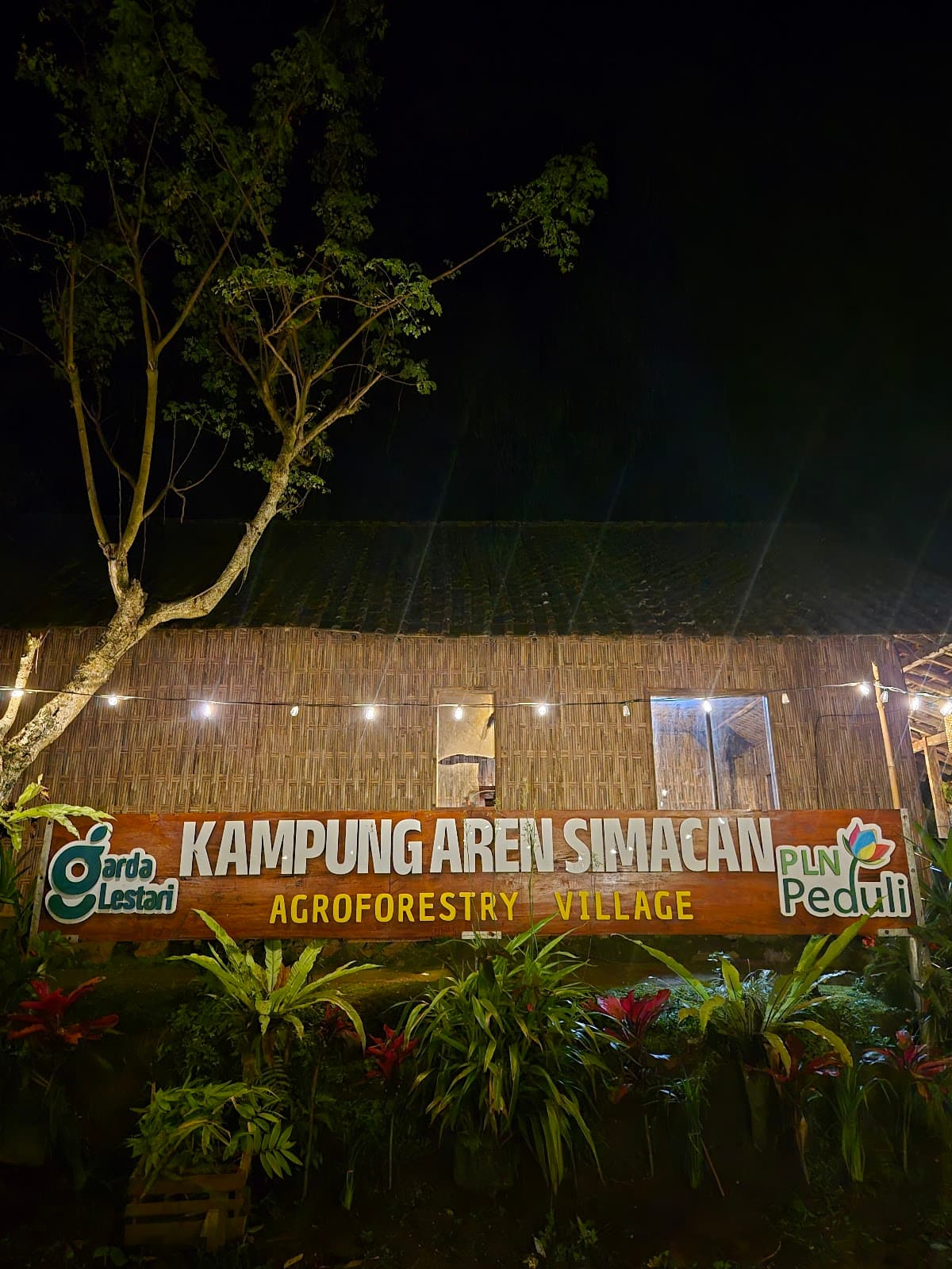 Papan nama Kampung Aren Simacan menyala di malam hari saat gerimis, dengan dinding bambu di belakangnya
