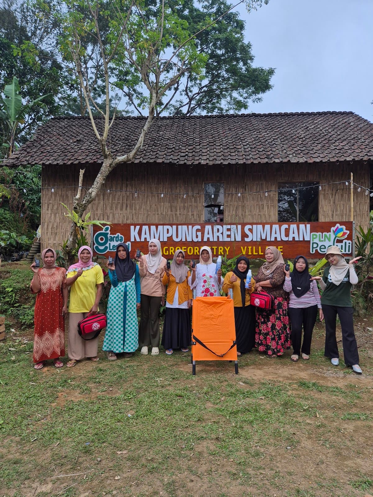 Ibu-Ibu Srikandi Kampung Aren Simacan memegang HT, tas P3K, alat pemadam, dan tandu lipat di depan papan nama kampung
