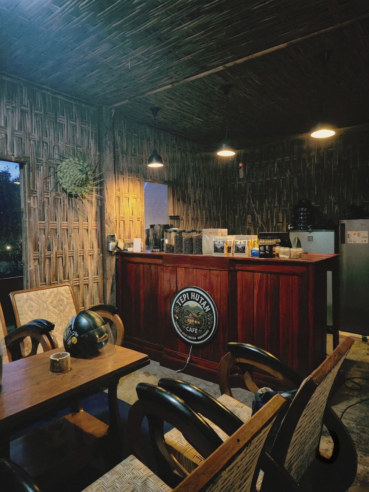 Interior Tepi Hutan Café dengan dinding dan langit-langit anyaman bambu, meja kayu, kursi rotan, dan bar berlogo café